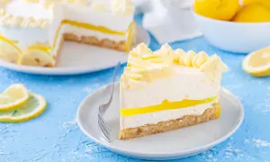 Una fetta di torta fredda al limone, con strati cremosi e una gelatina gialla brillante, decorata con ciuffi di panna e fette di limone.
