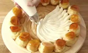 Una mano decora una torta con crema chiboust, creando un motivo a ventaglio. Piccoli bignè caramellati circondano la base.