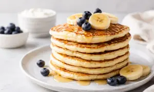 Pila di pancake proteici guarniti con mirtilli e banane, inzuppati nello sciroppo.