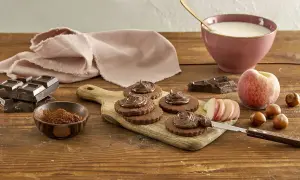 Biscotti senza burro al cacao, guarniti con crema al cioccolato, su tagliere di legno. Ingredienti: cioccolato, nocciole, pesca, latte.