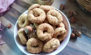 Ciambelline al vino cosparse di zucchero, con nocciole e un drappo rosa sullo sfondo.