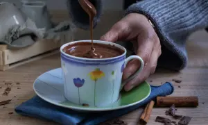 Una mano versa cioccolata calda in una tazza decorata con fiori. Cannella e scaglie di cioccolato completano la scena.