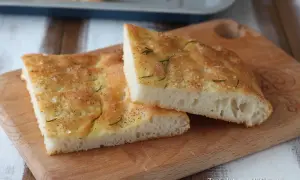 Focaccia senza glutine dorata e fragrante, condita con sale grosso e rosmarino, su un tagliere di legno.