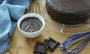 Torta ricoperta di lucida ganache cioccolato per copertura, con ciotolina di ganache, cioccolato fondente e frusta blu.