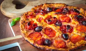 Focaccia pugliese dorata con pomodorini succosi e olive nere, guarnita con basilico fresco su tagliere di legno.