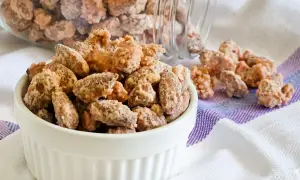 Un bianco contenitore pieno di mandorle pralinate, con altre sparse su un tovagliolo a righe.