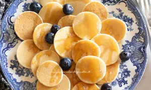 Mini pancake ricetta: tanti piccoli pancake dorati, guarniti con mirtilli freschi e sciroppo d'acero, serviti su un piatto decorato.