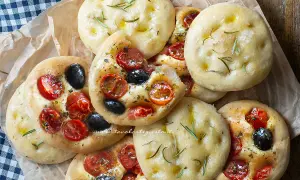Focaccine in padella, alcune condite con pomodorini e olive nere, altre con rosmarino e olio.