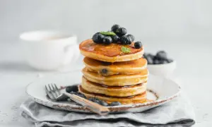 Pila di pancake con farina di riso, guarniti con mirtilli freschi e sciroppo d'acero, su un piatto rustico con una tazza di caffè sullo sfondo.