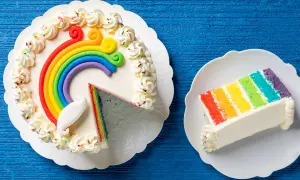 Torta arcobaleno con strati colorati e decorazioni di panna. Una fetta è già servita su un piattino.