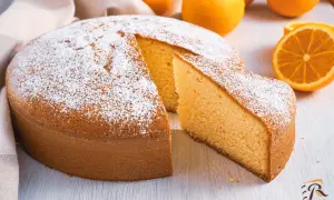 Torta soffice all'arancia, ricetta della nonna, cosparsa di zucchero a velo, con fette d'arancia sullo sfondo.