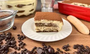 Una fetta di tiramisù con uova pastorizzate, cosparso di cacao, su un piatto bianco. Intorno, chicchi di caffè, palline di cioccolato e un savoiardo.