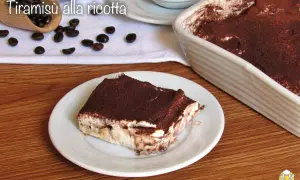 Una fetta di tiramisù alla ricotta, cosparso di cacao, su un piatto bianco, con chicchi di caffè e una teglia sullo sfondo.