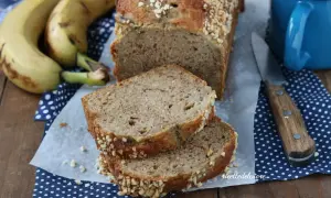 Fetta di banana bread senza burro, soffice e profumato, con crosta croccante di frutta secca. Accanto, banane mature e una tazza blu.