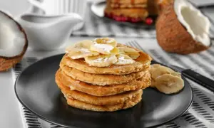 Pancakes con farina di cocco, guarniti con banane a fette e sciroppo, su un piatto nero. A lato, un cocco aperto.