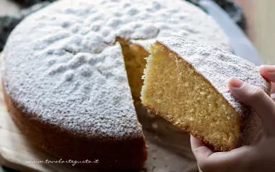 Una fetta di torta margherita soffice e profumata, cosparsa di zucchero a velo, viene sollevata da una mano.