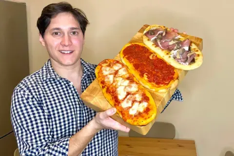 Uomo sorridente mostra tre lingue di pizza senza lievito: una margherita, una al pomodoro e una con prosciutto e funghi.