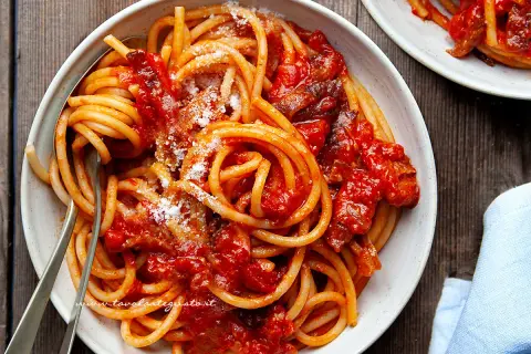 Un piatto di bucatini all'amatriciana ricetta, conditi con sugo rosso e guanciale croccante, spolverati di pecorino.