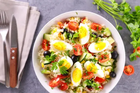 Una gustosa insalata di riso con uova sode, pomodorini, olive, mais e cetrioli, guarnita con prezzemolo fresco.