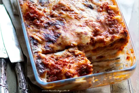 Lasagna al ragù appena sfornata, dorata e filante, in una teglia di vetro, con posate d'argento a lato.