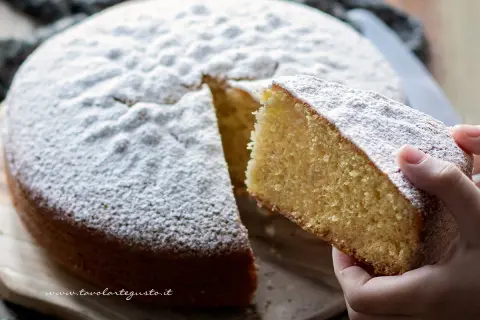 Una fetta di torta margherita soffice e profumata, cosparsa di zucchero a velo, viene sollevata da una mano.