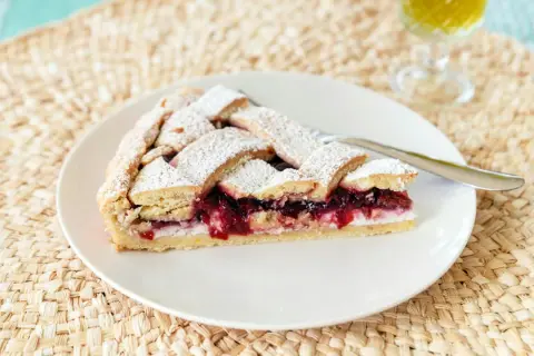 Una fetta di crostata di ricotta con confettura di ciliegie, cosparsa di zucchero a velo, su un piatto bianco.