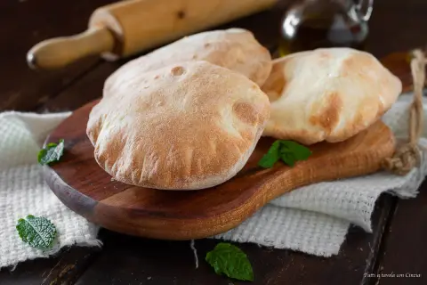 Tre soffici pane arabo dorati su un tagliere di legno, guarniti con foglie di menta fresca.