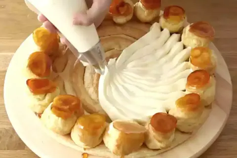 Una mano decora una torta con crema chiboust, creando un motivo a ventaglio. Piccoli bignè caramellati circondano la base.