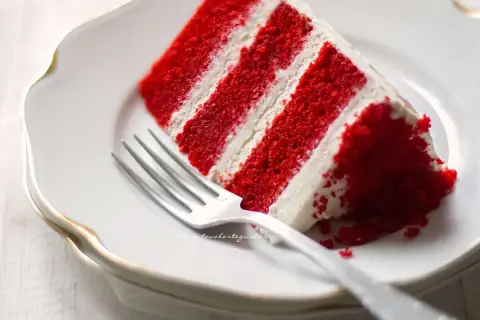 Una fetta di torta red velvet, stratificata con crema bianca, su un piatto bianco con una forchetta.