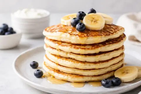 Pila di pancake proteici guarniti con mirtilli e banane, inzuppati nello sciroppo.