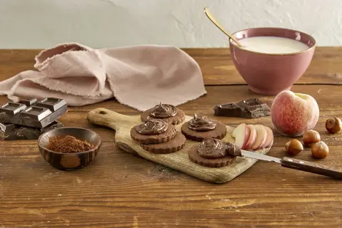 Biscotti senza burro al cacao, guarniti con crema al cioccolato, su tagliere di legno. Ingredienti: cioccolato, nocciole, pesca, latte.