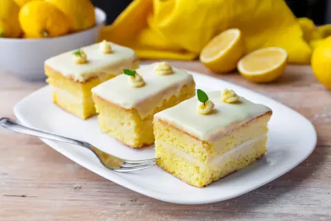 Tre fette di torta al limone e cioccolato bianco, guarnite con glassa e foglioline verdi, su un piatto bianco con limoni sullo sfondo.