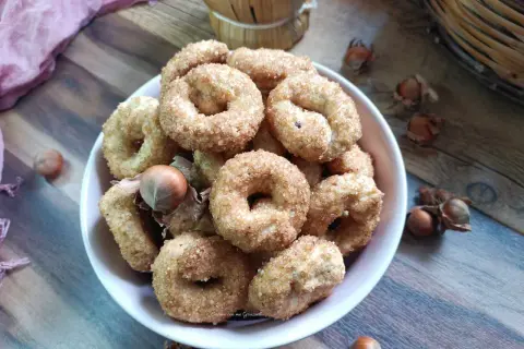 Ciambelline al vino cosparse di zucchero, con nocciole e un drappo rosa sullo sfondo.