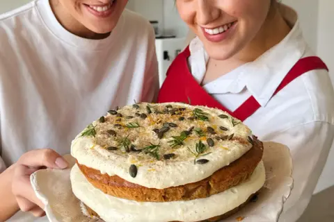 Due persone sorridono mostrando una torta a strati decorata con glassa, semi di zucca e scorza d'arancia. La ricetta della carrot cake è un successo!