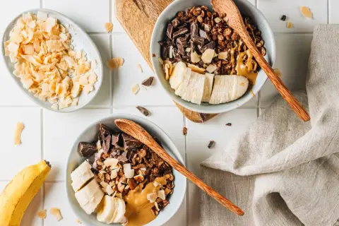 Due ciotole con granola fatta in casa, banana a fette, cioccolato fondente e burro d'arachidi. A lato, scaglie di cocco tostato.