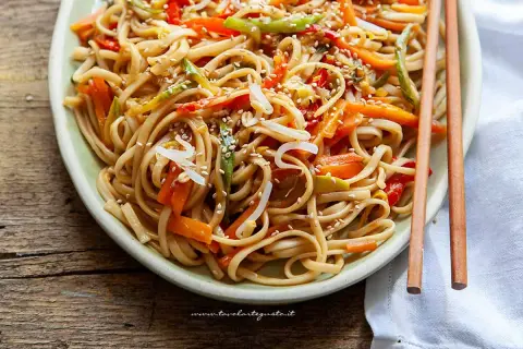 Un piatto invitante di spaghetti di riso saltati con verdure colorate e semi di sesamo, pronti per essere gustati con le bacchette.