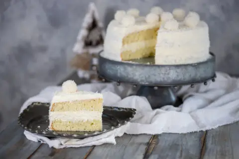 Una fetta di torta Raffaello, soffice e ricoperta di cocco, su un piatto decorato. Sullo sfondo, la torta intera su un'alzata.