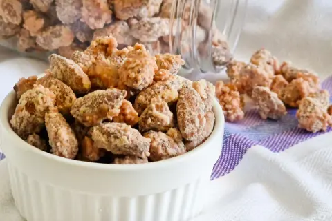 Un bianco contenitore pieno di mandorle pralinate, con altre sparse su un tovagliolo a righe.