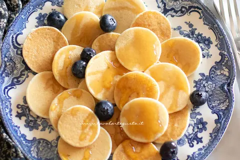 Mini pancake ricetta: tanti piccoli pancake dorati, guarniti con mirtilli freschi e sciroppo d'acero, serviti su un piatto decorato.