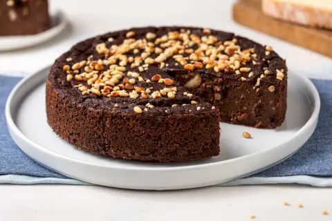 Torta paesana al cioccolato, ricca e umida, guarnita con pinoli tostati. Una fetta è stata già servita, invitando a gustare questo dolce rustico.