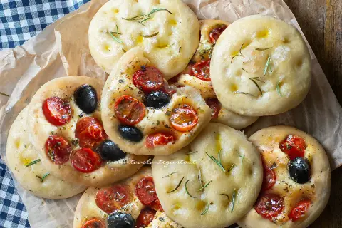 Focaccine in padella, alcune condite con pomodorini e olive nere, altre con rosmarino e olio.