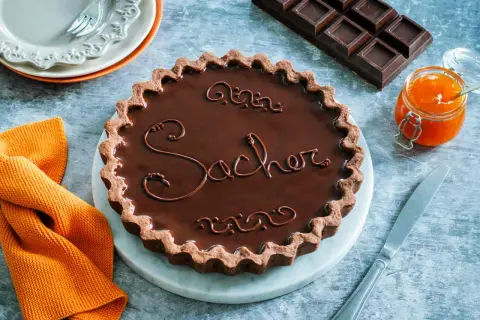 Una crostata Sacher lucida e invitante, con decorazioni in cioccolato e un vasetto di marmellata d'arancia accanto.