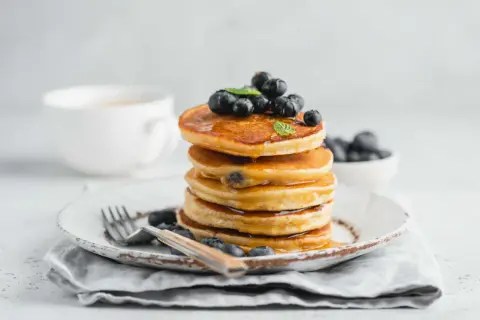Pila di pancake con farina di riso, guarniti con mirtilli freschi e sciroppo d'acero, su un piatto rustico con una tazza di caffè sullo sfondo.
