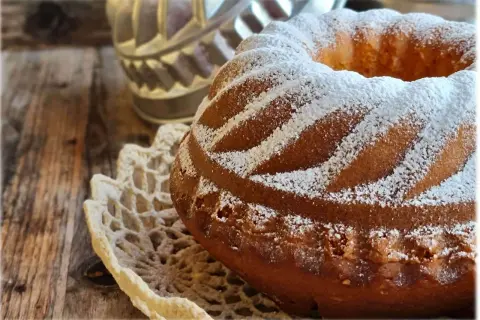 Ciambellone soffice come una nuvola, cosparso di zucchero a velo, adagiato su un centrino di pizzo, su un tavolo di legno rustico.