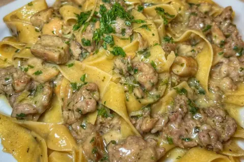 Pappardelle cremose con salsiccia e funghi, guarnite con prezzemolo fresco. Un piatto ricco e saporito.