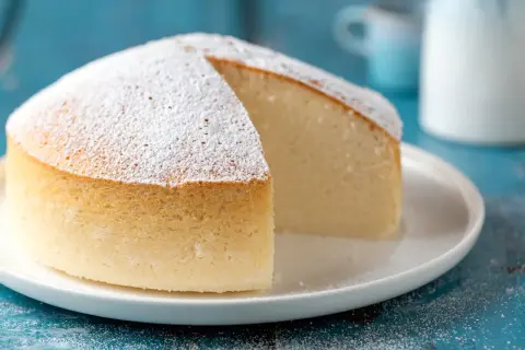 Una soffice torta nuvola, cosparsa di zucchero a velo, su un piatto bianco. La sua consistenza leggera invita a un morso goloso.