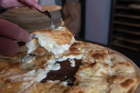 Una fetta di focaccia di Recco ricetta, filante e dorata, viene sollevata con una forchetta.