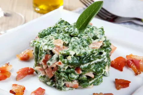 Spätzle verdi cremosi con pezzetti di speck croccante, guarniti con una foglia di salvia. Un piatto invitante.