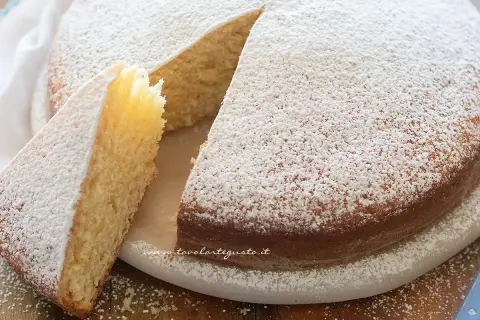 Una soffice torta senza uova, cosparsa di zucchero a velo, con una fetta tagliata che ne mostra l'interno chiaro e profumato.