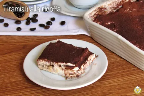 Una fetta di tiramisù alla ricotta, cosparso di cacao, su un piatto bianco, con chicchi di caffè e una teglia sullo sfondo.